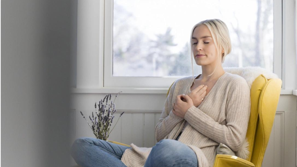Mulher loira sentada em uma poltrona amarela com os olhos fechados e mãos no peito, praticando meditação e controle da respiração. Ao fundo, uma janela e um buquê de lavanda, transmitindo calma e bem-estar