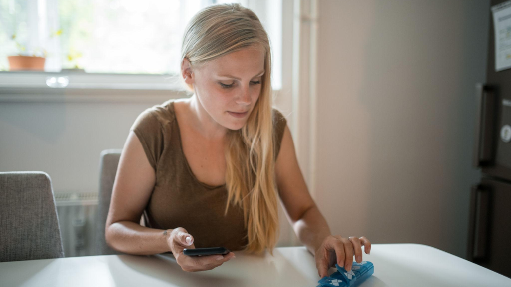 Jovem loira utiliza um smartphone para fazer compras online de um organizador de comprimidos azul, destacando o papel da tecnologia na gestão da saúde doméstica. A interface de compras com texto verde 'EM ESTOQUE: 5' está visível.